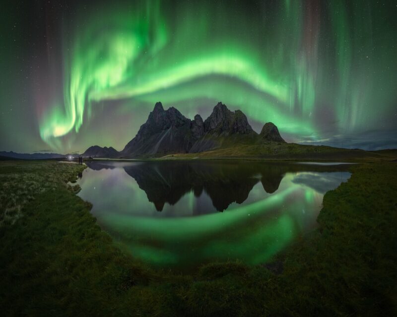 Swirling green aurora over sharp mountains with a reflection in the lake.