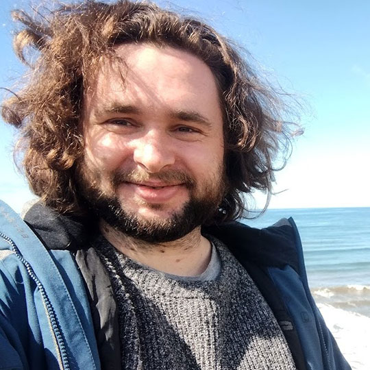 Young, long-haired, beardedd man standing by the ocean.