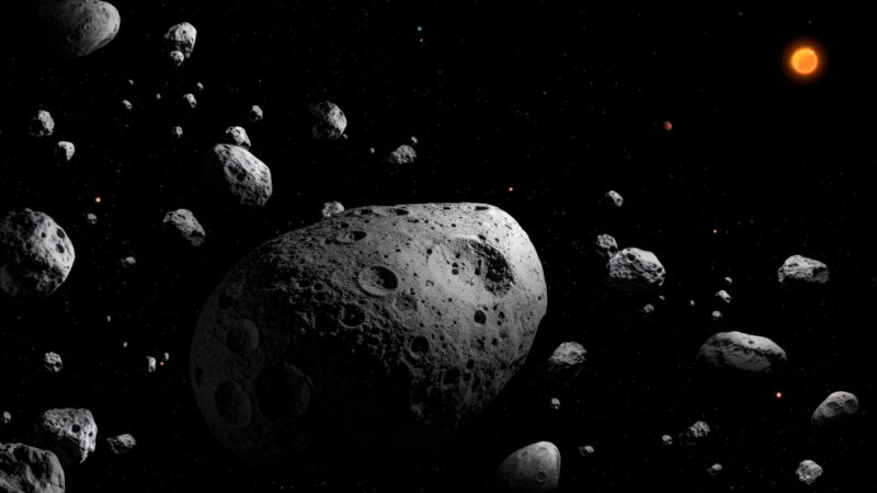 Irregular, cratered gray asteroid in a field of other asteroids. Sun and tiny Jupiter in background.