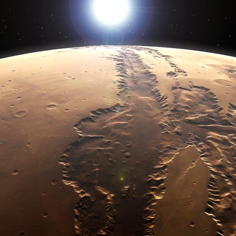Huge canyon system on a brownish-red planet viewed from space, with a bright sun in the distance.