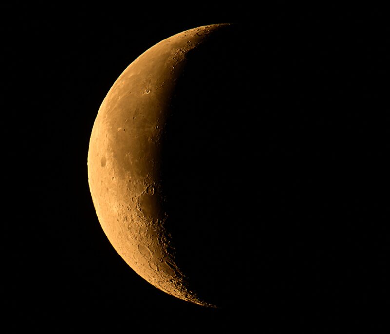 A closeup of an orange-colored, sideways-looking waning crescent moon.