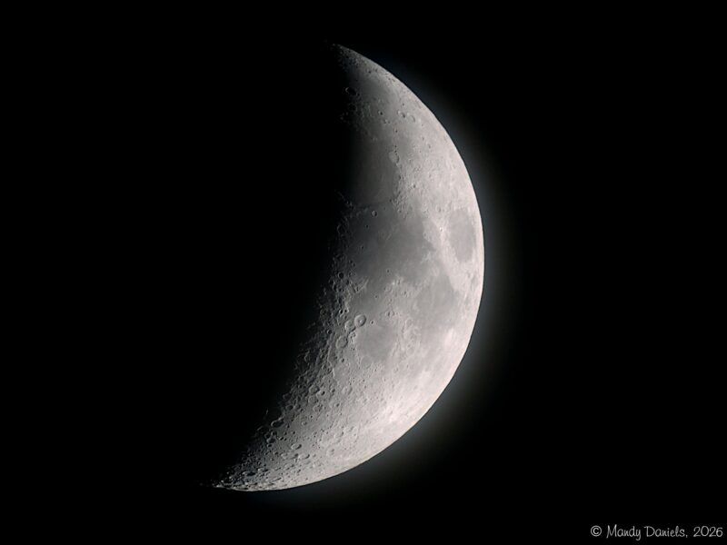 A thick crescent moon on a black background, mostly gray with darker blotches and many craters.