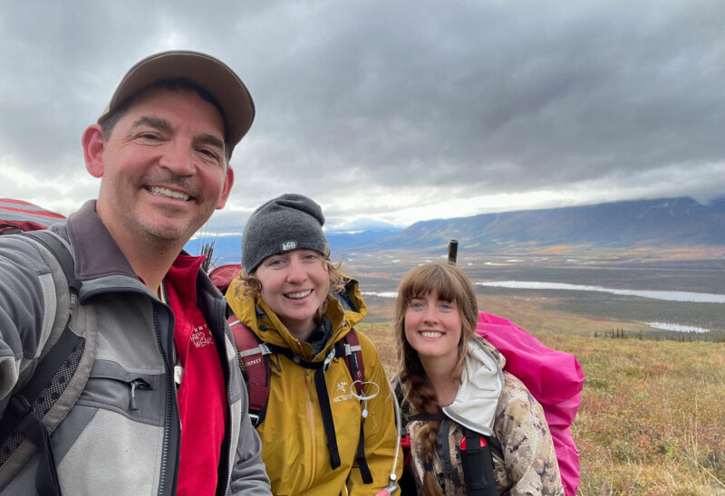 A man and 2 women similing in a landscape with low vegetation and distant hills under a cloudy sky.