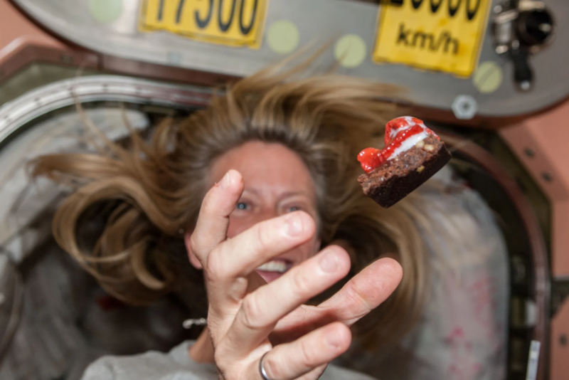 Inside a spacecraft, a smiling woman with her hair floating reaches for a floating brownie with toppings.