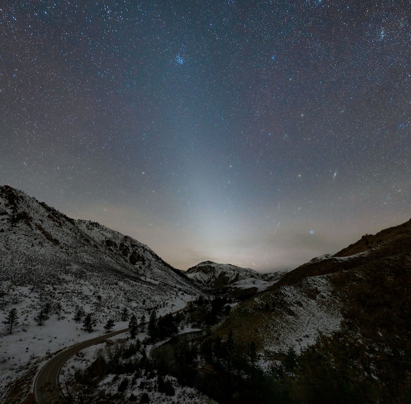 A pyramid white cone of light slanting left on a starry sky and over a dark winding road surrounded by snowy mountains.