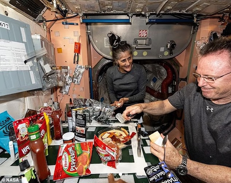 In a cramped spacecraft, 2 smiling astronauts at a table with a wide variety of labeled packets.
