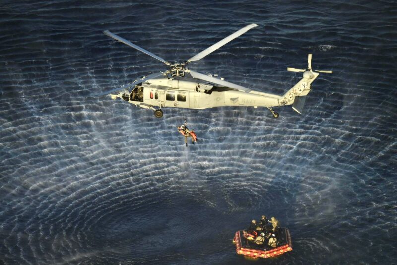 Helicopter hovering above raft floating in the ocean, with humans aboard. One person is being lifted toward the helicopter.