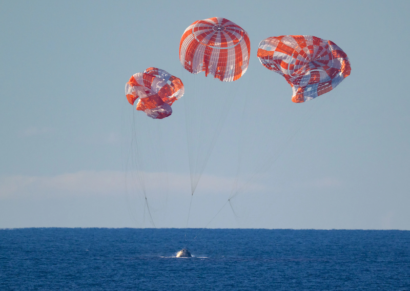 A space capsule splashes into a wide blue sea, with red and white parachutes beginning to collapse above.