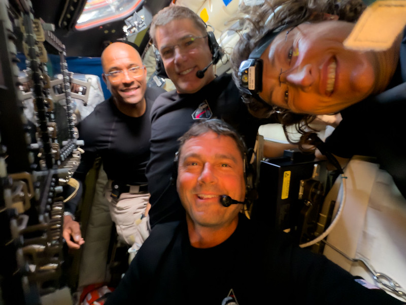 Four smiling astronauts get close together for a pic. Two are wearing mics. You can see equipment on the left.