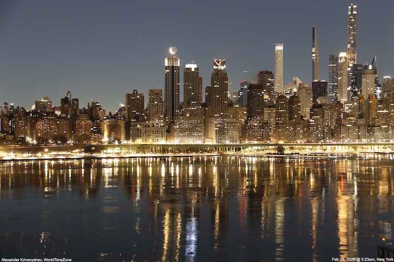 Manhattan skyline in dark twilight reflecting in water with crescent moon glowing with earthshine over a building.