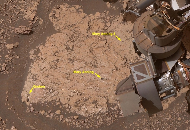Flattish brown rock seen from above. 3 small holes are labeled and part of a robotic arm is on the right side.