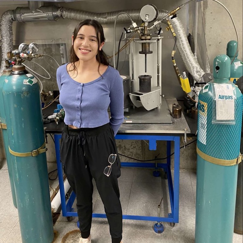 Smiling young woman with blue shirt and black pants standing in front of a mechanical apparatus in a lab.