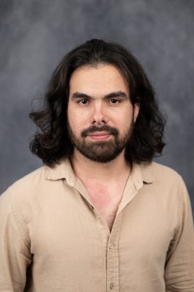 Light skinned male with dark shoulder length hair and beard in a light brown shirt.