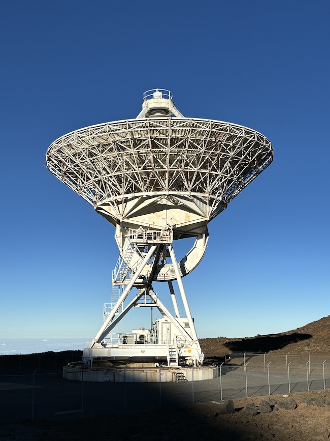 A large dish-shaped antenna on a barren brown mountaintop under a deep blue sky. A fence in the foreground.