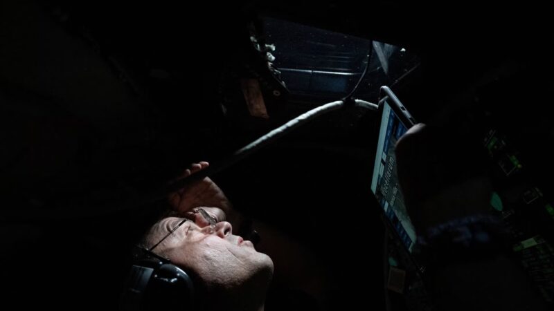 Profile of a man in a darkened room, peering upward through machinery.