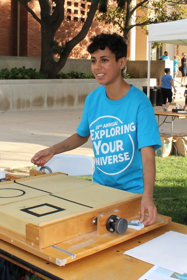 Young woman with short dark hair standing at a table outdoors with a flat, square wooden box on it.