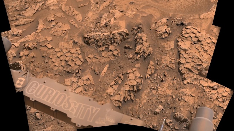 Brownish sandy terrain with rocks covered in what look like reptile scales. A part of a machine with the word Curiosity on it is in the foreground.