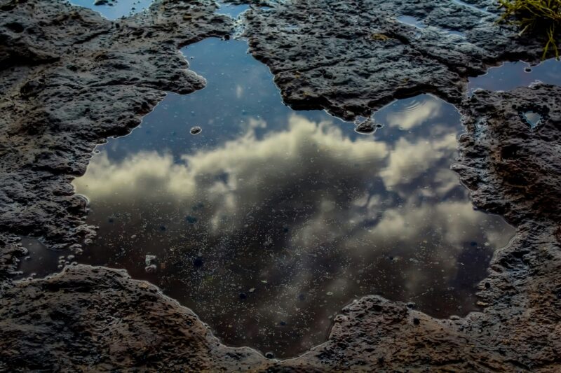 A puddle rests on brown earth, its surface reflecting the clouds above.