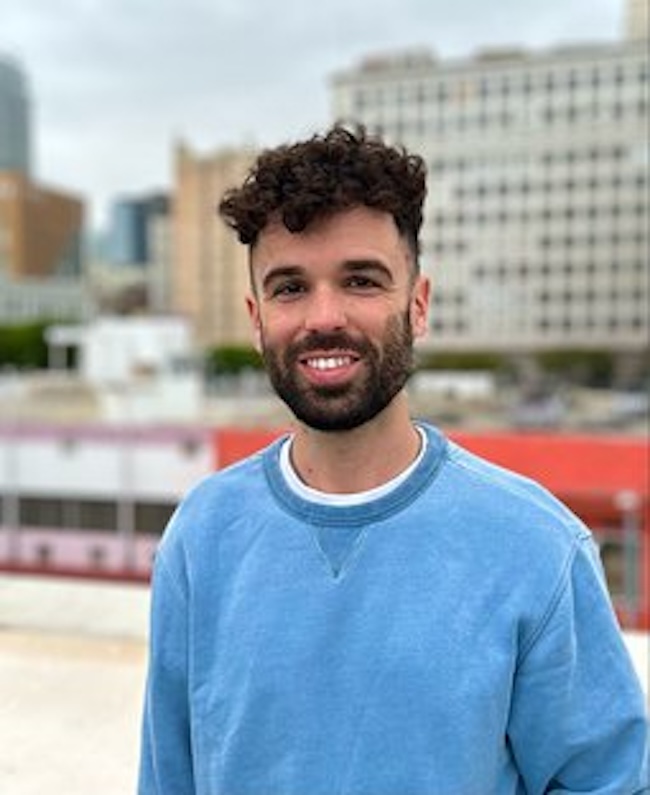 Smiling man with dark, curly hair and beard, wearing a blue long-sleeved shirt.