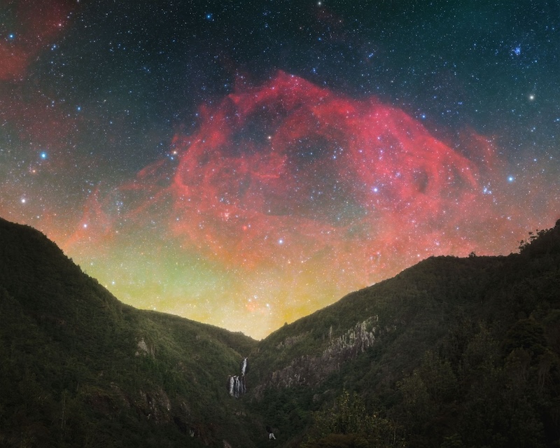 Starry night sky showing large reddish nebula, lots of stars and a greenish airglow above the mountains.