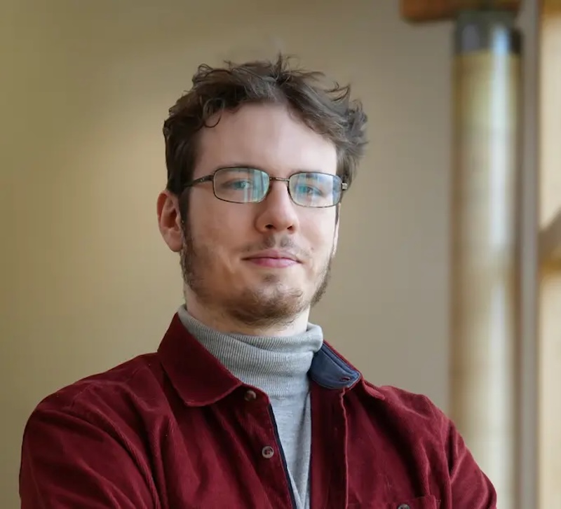Smiling man with tousled hair and eyeglasses, wearing a brown jacket.