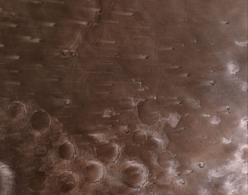 Reddish-brown ground with many craters with lighter streaks, all in one direction, from them.