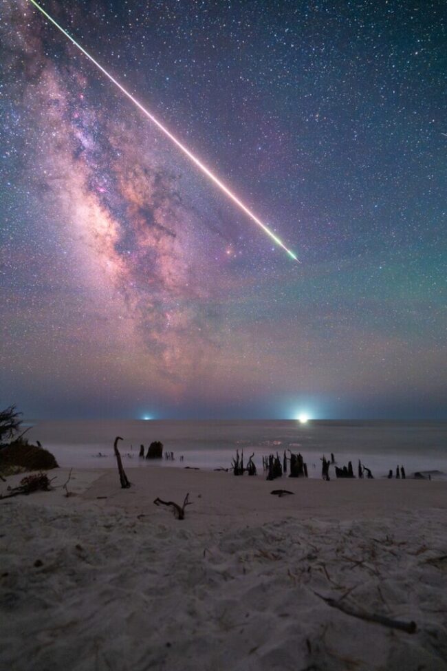 Beach scene with the Milky Way stretching upward and a fireball meteor running along the top edge.