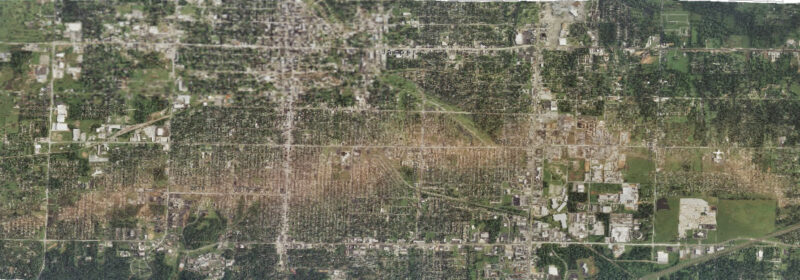 Satellite view of a city with a long, light brown swath across many blocks otherwise green with trees.