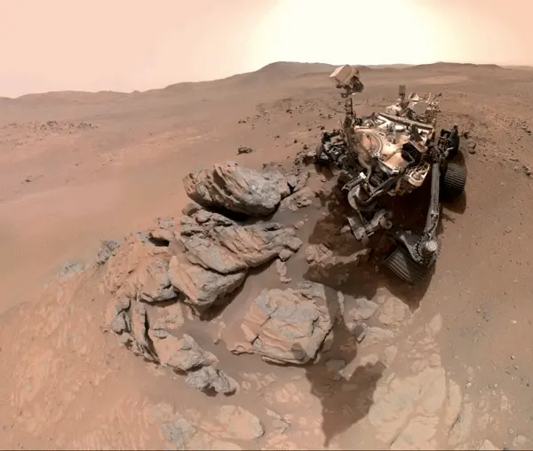 A complicated wheeled vehicle with a camera up on an arm, near a pile of boulders.