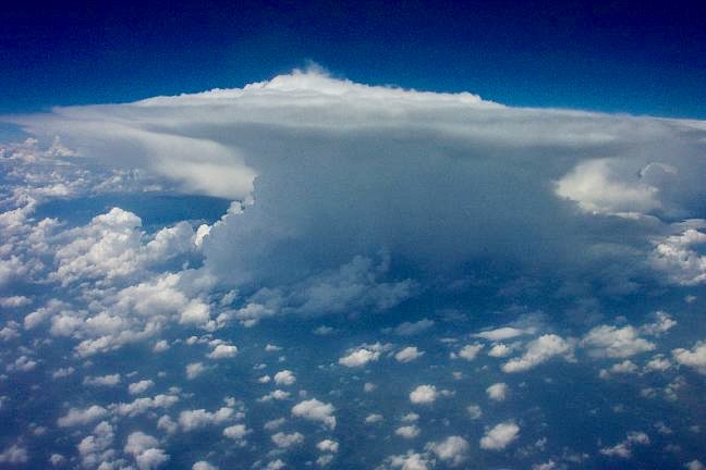 Gigantic-looking anvil shaped cloud, rising above a puffy cloud layer.