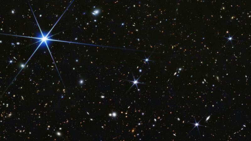 Aliens visit Earth: A field of a myriad tiny, distant galaxies and one bright star at upper left with spikes.