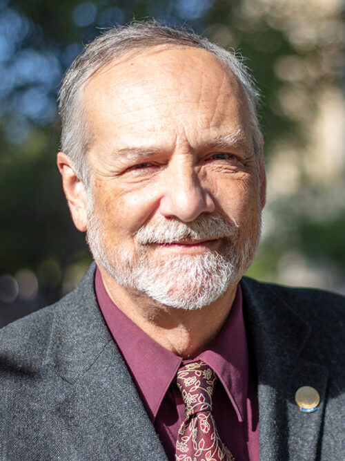 A man with gray hair and beard wearing a gray suit with mauve shirt.