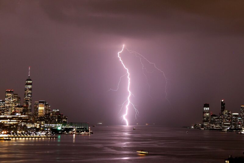 Cities can change the weather: Vertical lightning bolt strikes water between 2 large cities glittering with lights at night.