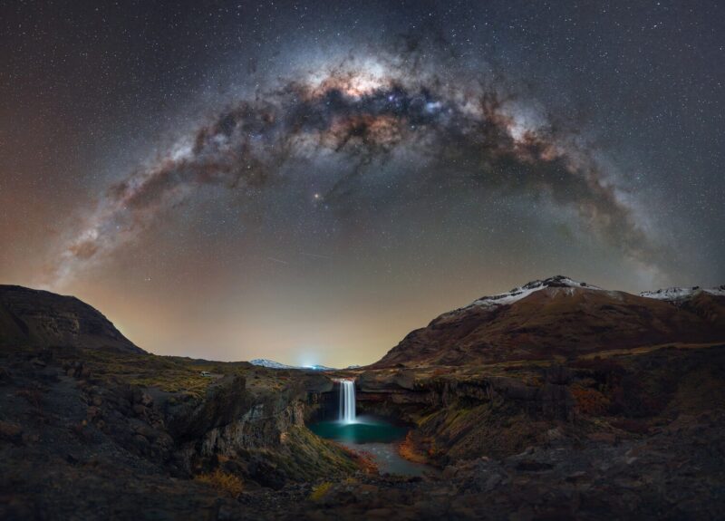 A Milky Way arches over landscape with a waterfall.