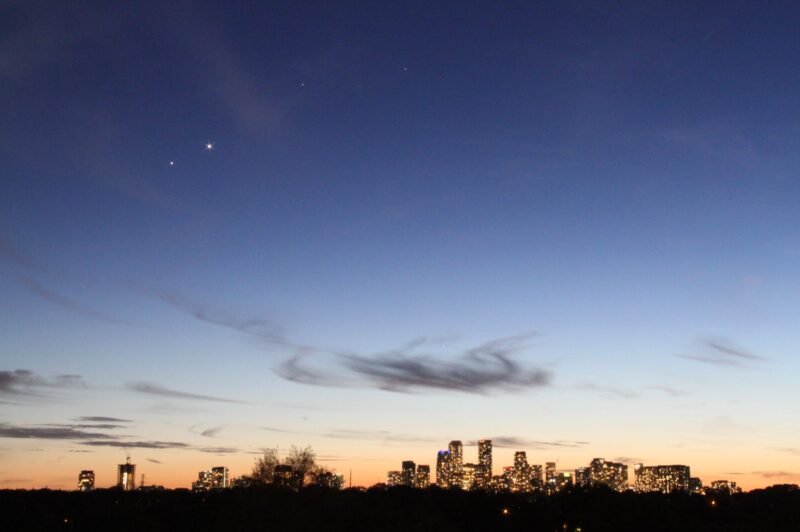 2 bright dots and 2 other fainter dots in the twilight sky above a glittering city skyline.