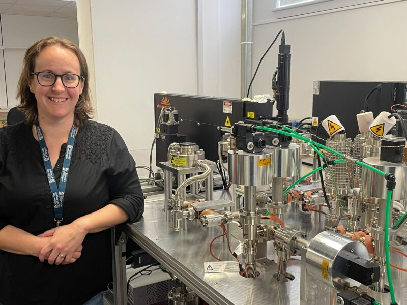 A woman wearing glasses and a lanyard stands next to testing equipment.