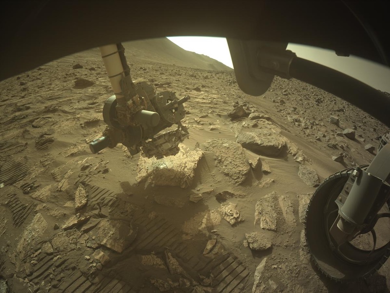 Carbon in Mars rocks: Brown, barren rocky terrain. A robotic arm and 1 wheel of a rover are visible.