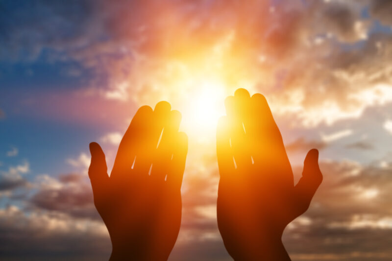 A sky with orange clouds and a low sun, with 2 hands cupping the sun.