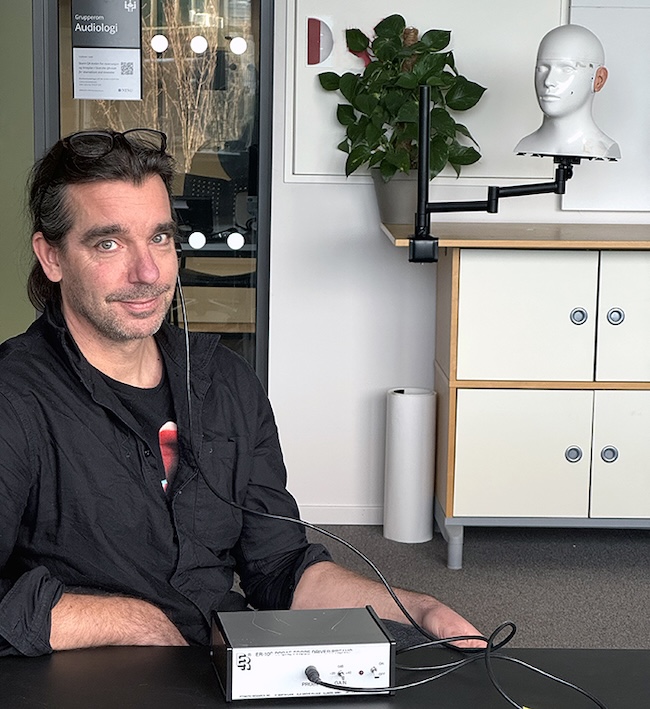 Man sitting at a desk with a wire going from a small aluminum box to his ear.