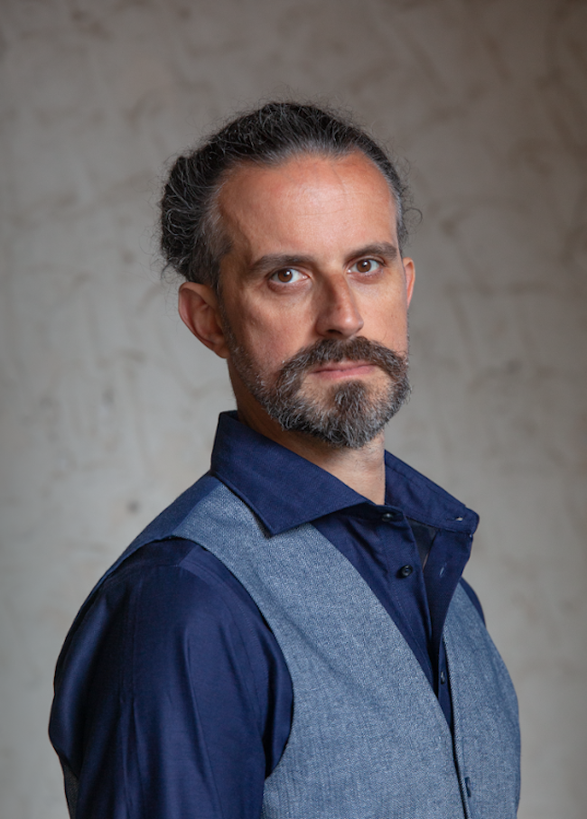 Serious-looking man with beard wearing a dark blue dress shirt and light blue vest.
