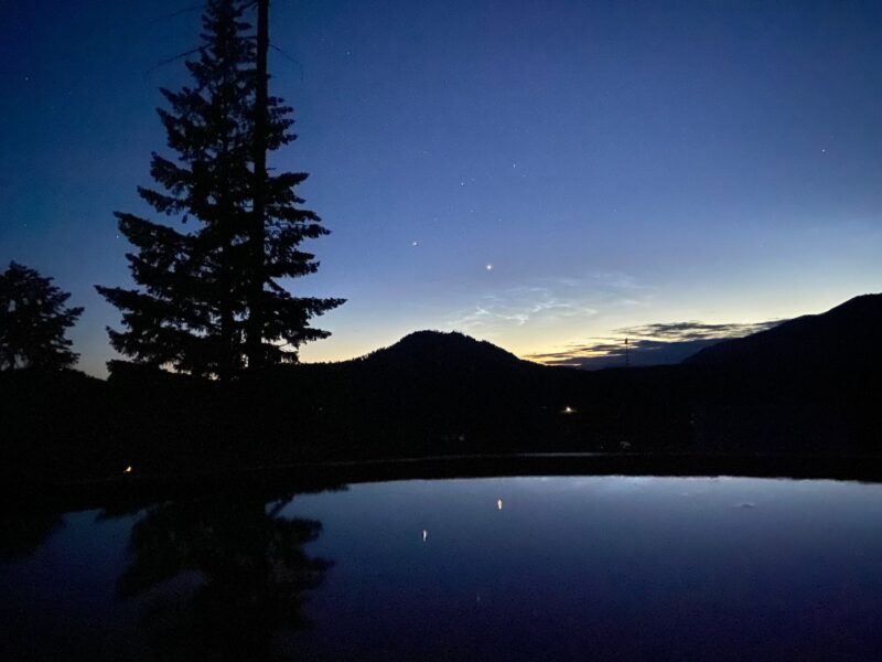 Bright dots in the sky reflected in the water below. Trees on the left and soft mountains in the distance.