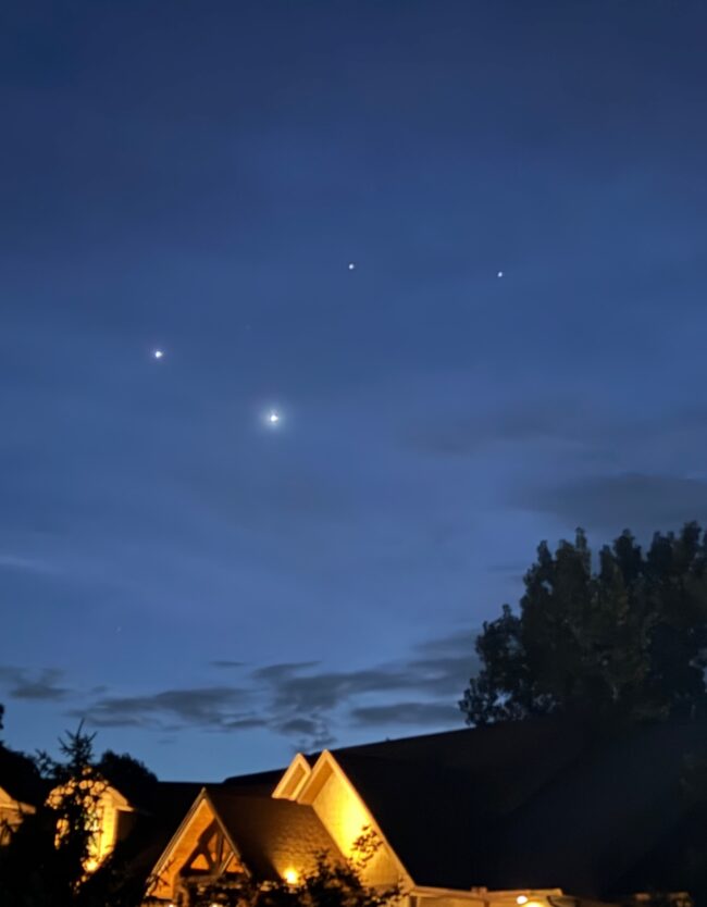 4 bright dots in a dark blue sky, with rooftops and trees below.