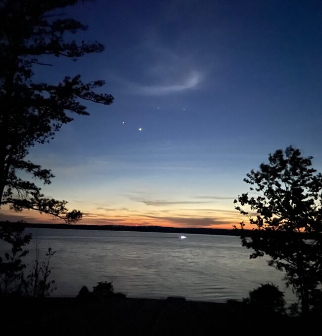 Dark blue sky and orange horizon over the water, with trees in the foreground. Four bright dots in the sky.
