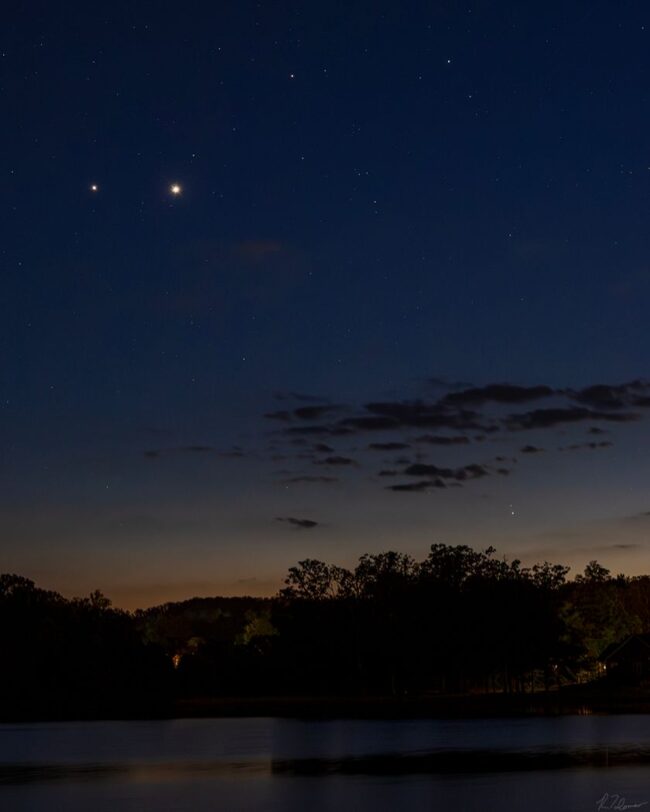 2 bright dots in a deep blue sky fading into orange on the horizon, above trees by a body of water.