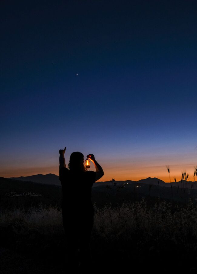 A person holding a lantern in one hand and pointing at the dark blue sky with the other.
