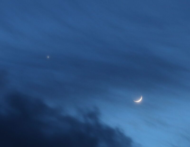 A crescent moon next to Venus and some clouds in a sky that is growing dark.