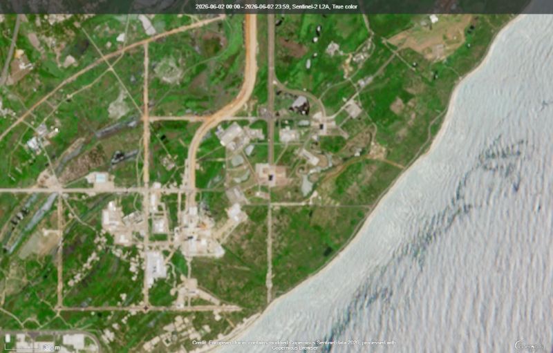 Water at lower right and green grass plus tan buildings, roads, and a launch pad near center.