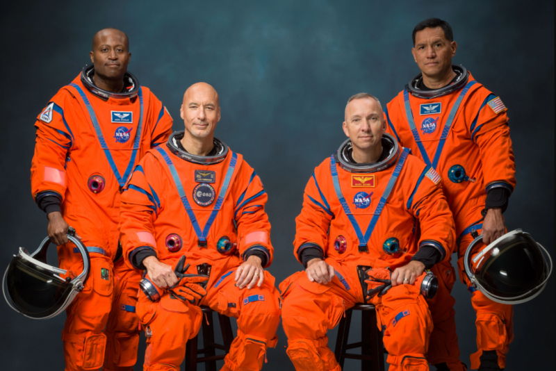 Four men in orange flight suits, 2 seated and 2 stsnding holding space helmets, posing for a portrait.