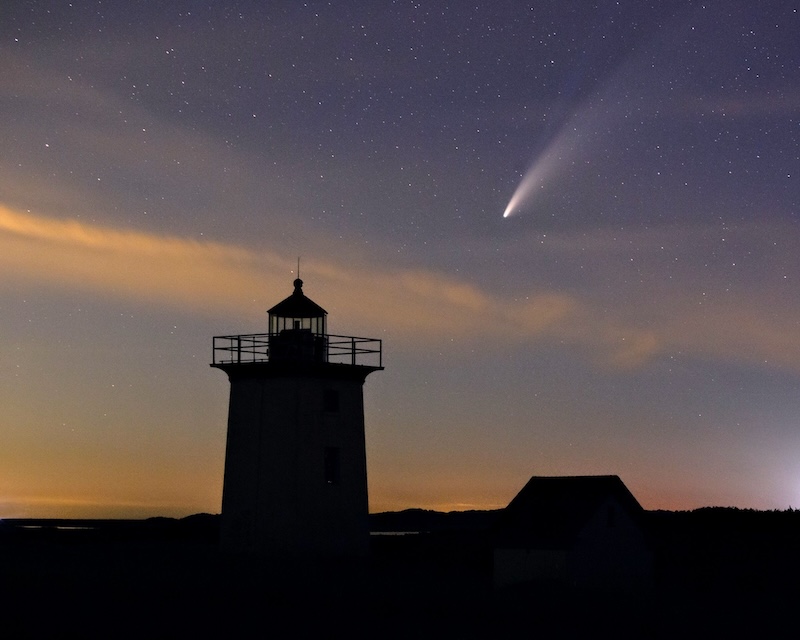 Bright comet in a starry sky above a rank of clouds. A lighthouse is silhouetted in the foreground.