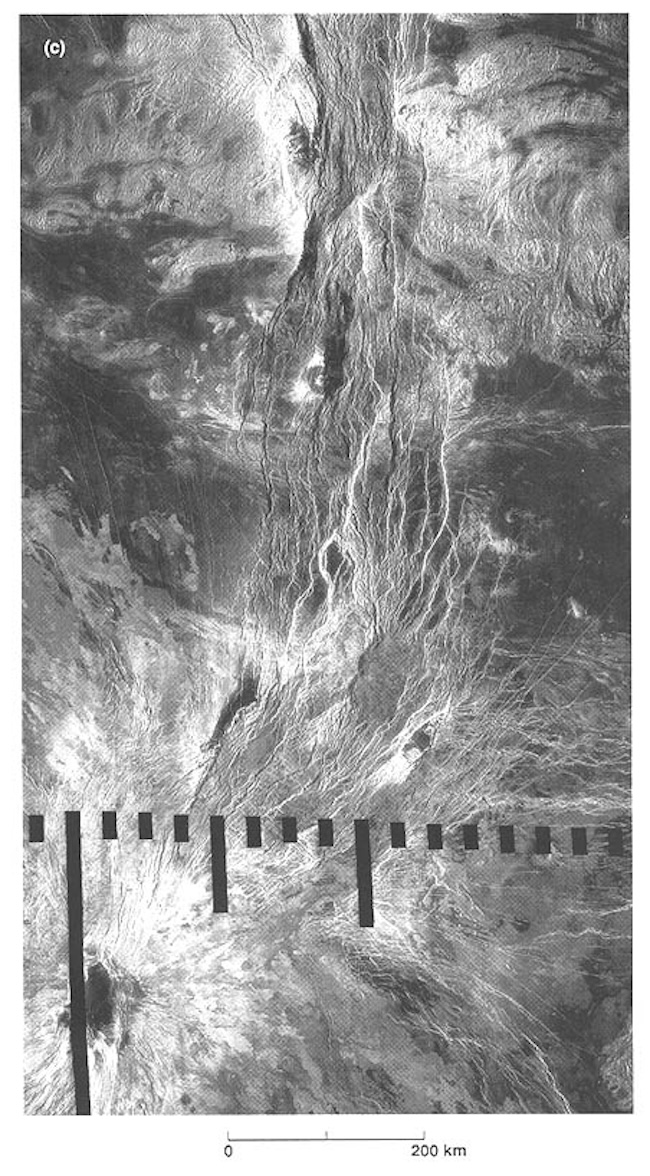 Black and white overhead image of a bright volcano with dark center in lower left. Many thin, bright lines are radiating from the volcano to the upper right.